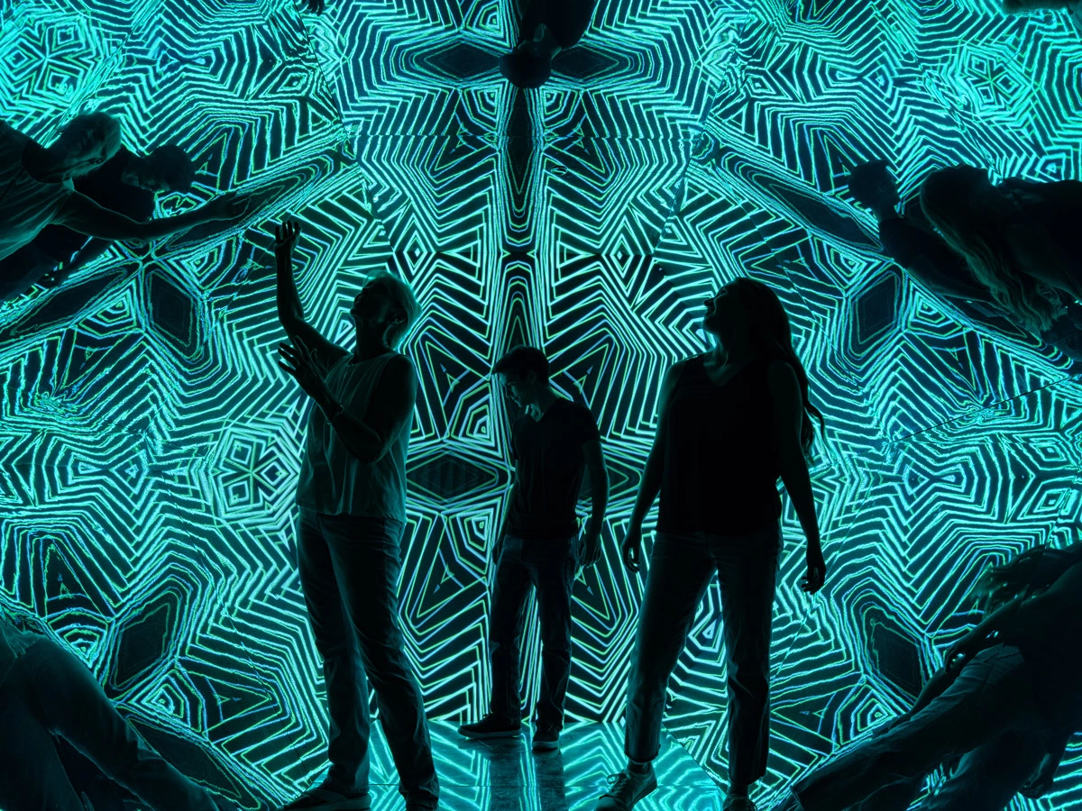 Three persons are standing in the dark of the Walk in Kaleidoscope at Museum of Illusions Las Vegas. Their outlines are seen in front of the exhibit's geometric turquoise and black pattern.