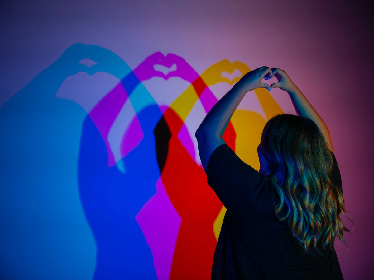 A woman forms a heart shape with her hands in front of a wall of the Color Room at Museum of Illusions Las Vegas. Her shadows on the wall are seen in various colours, from blue to magenta, red, and yellow.