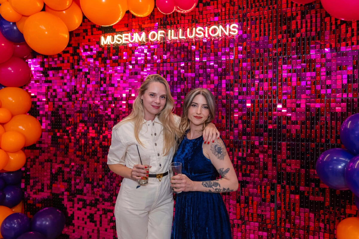 Two young women are posing for a photo in front of a magenta glittery wall with a Museum of Illusions sign.