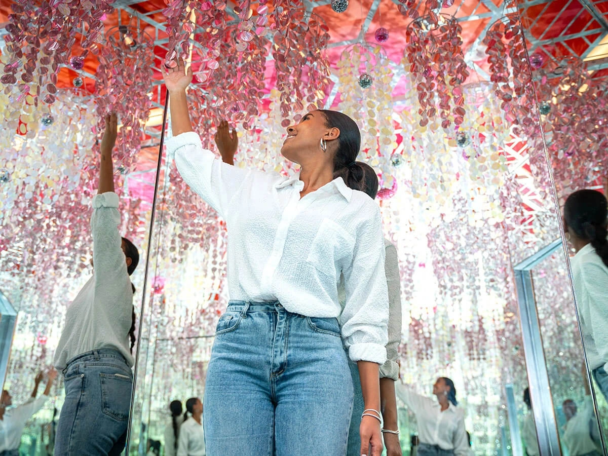 A smiling woman is reaching her hand towards the glittery cascades at Museum of Illusions Las Vegas Infinity Room. Her infinite reflections are seen in the mirrors on the walls.