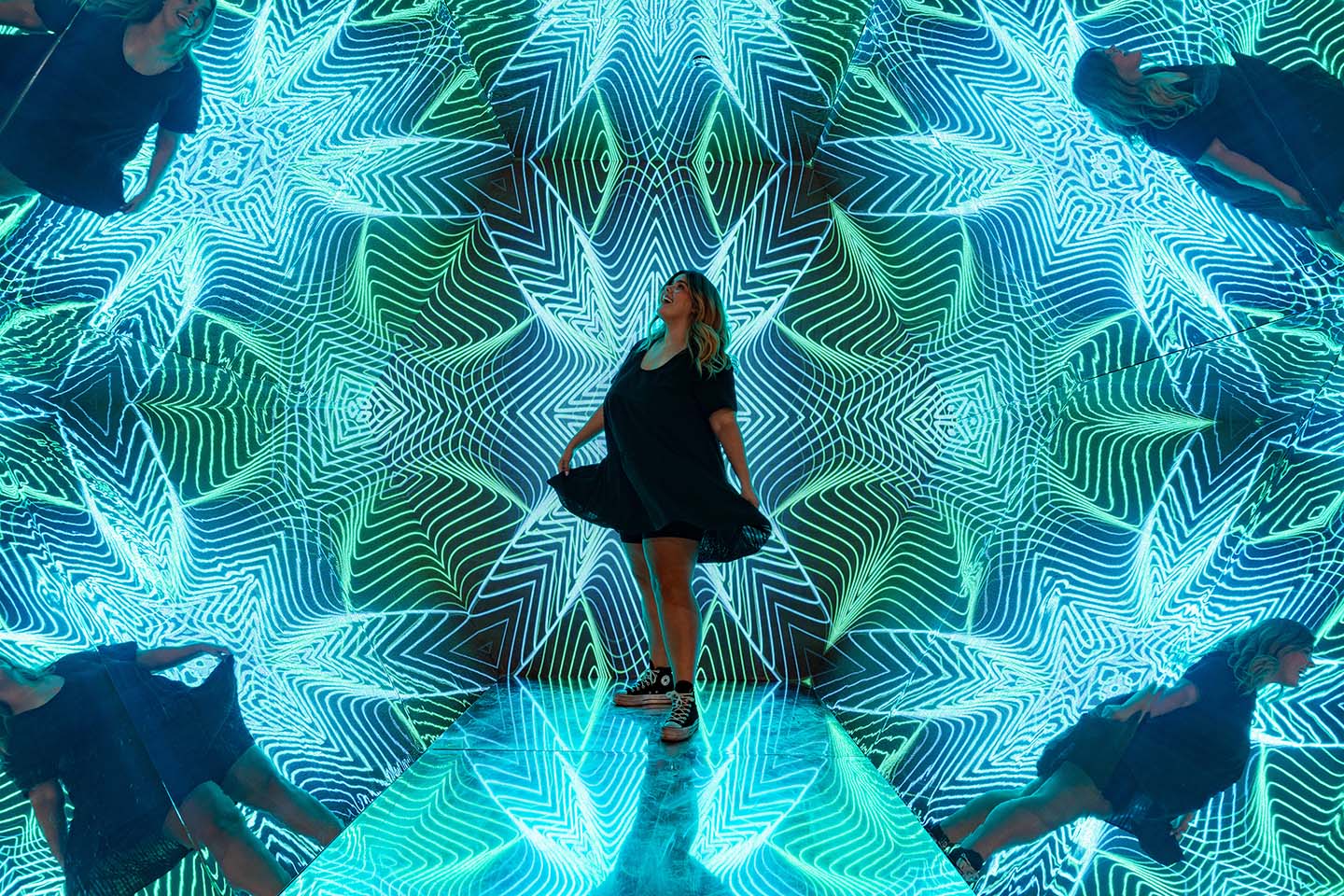 A woman in a dress is standing in the Walk in Kaleidoscope at Museum of Illusions Las Vegas. She is surrounded by the turquoise-tinted geometrical pattern.